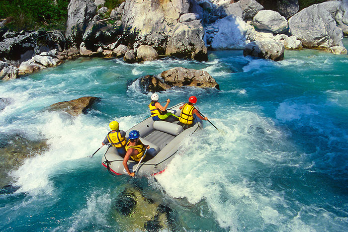 Group of four people white-water rafting through rapids in a blue river, surrounded by rocks.