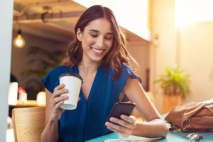 Woman smiling at her phone while holding a cup.