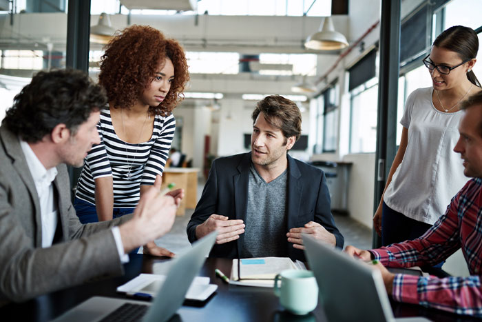 A group of five people have a discussion around a table in a bright office space with laptops and notebooks.