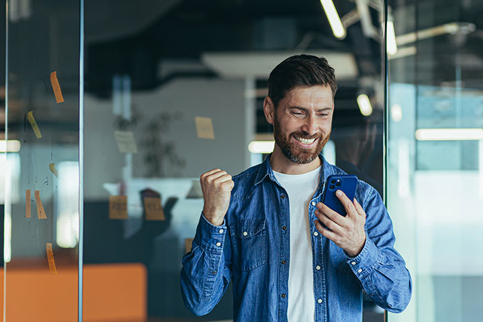 A man in a blue shirt smiling and holding a phone in an office setting.